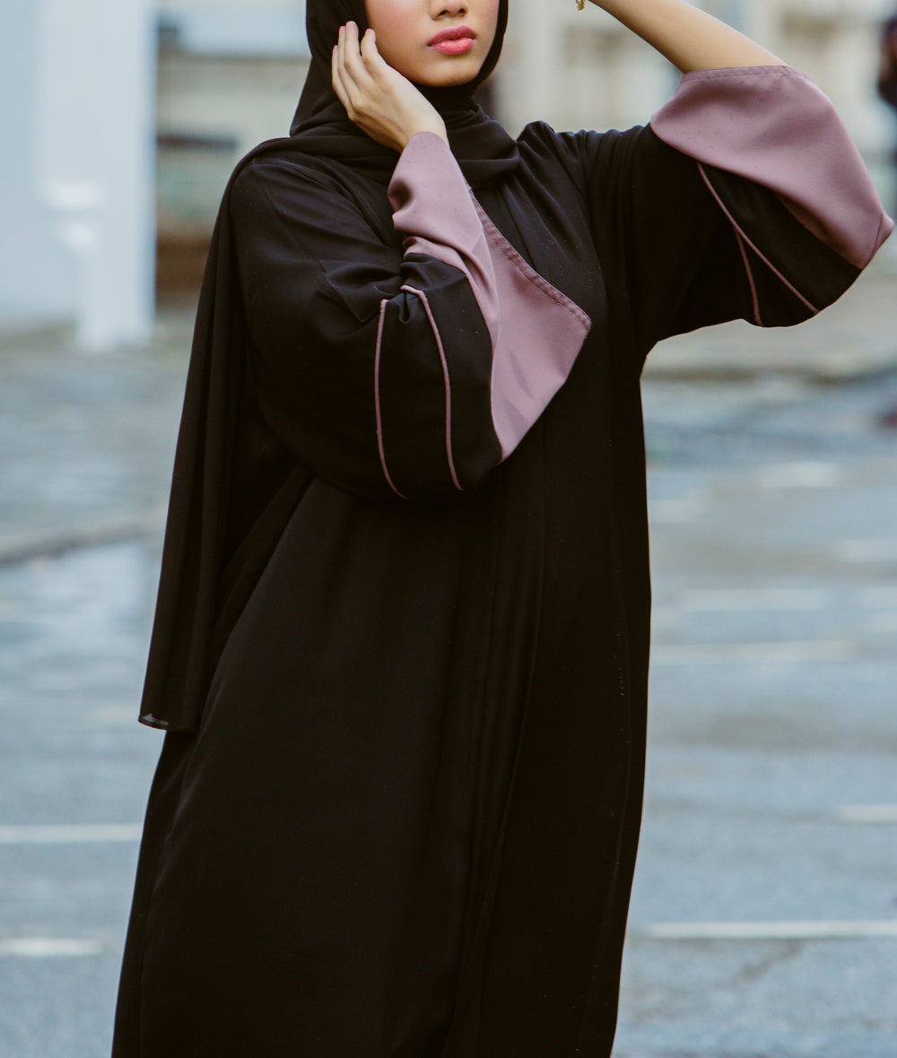 A woman wearing a minimalist black abaya dress with wide mauve contrast sleeves and delicate piping from the latest abaya collection 2026.