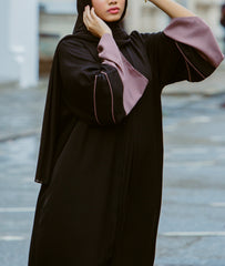A woman wearing a minimalist black abaya dress with wide mauve contrast sleeves and delicate piping from the latest abaya collection 2026.
