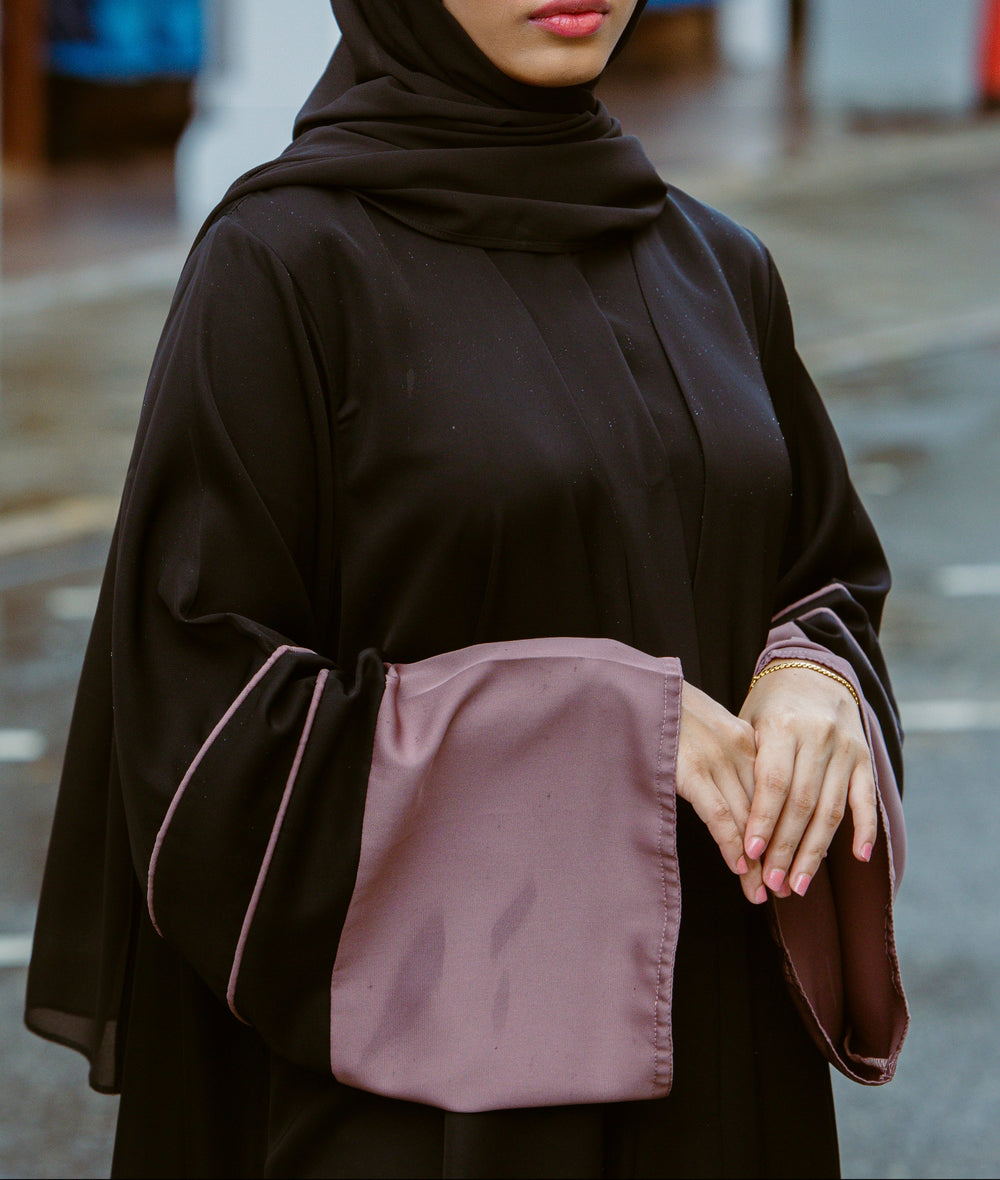 A woman wearing a minimalist black abaya dress with wide mauve contrast sleeves and delicate piping from the latest abaya collection 2026.