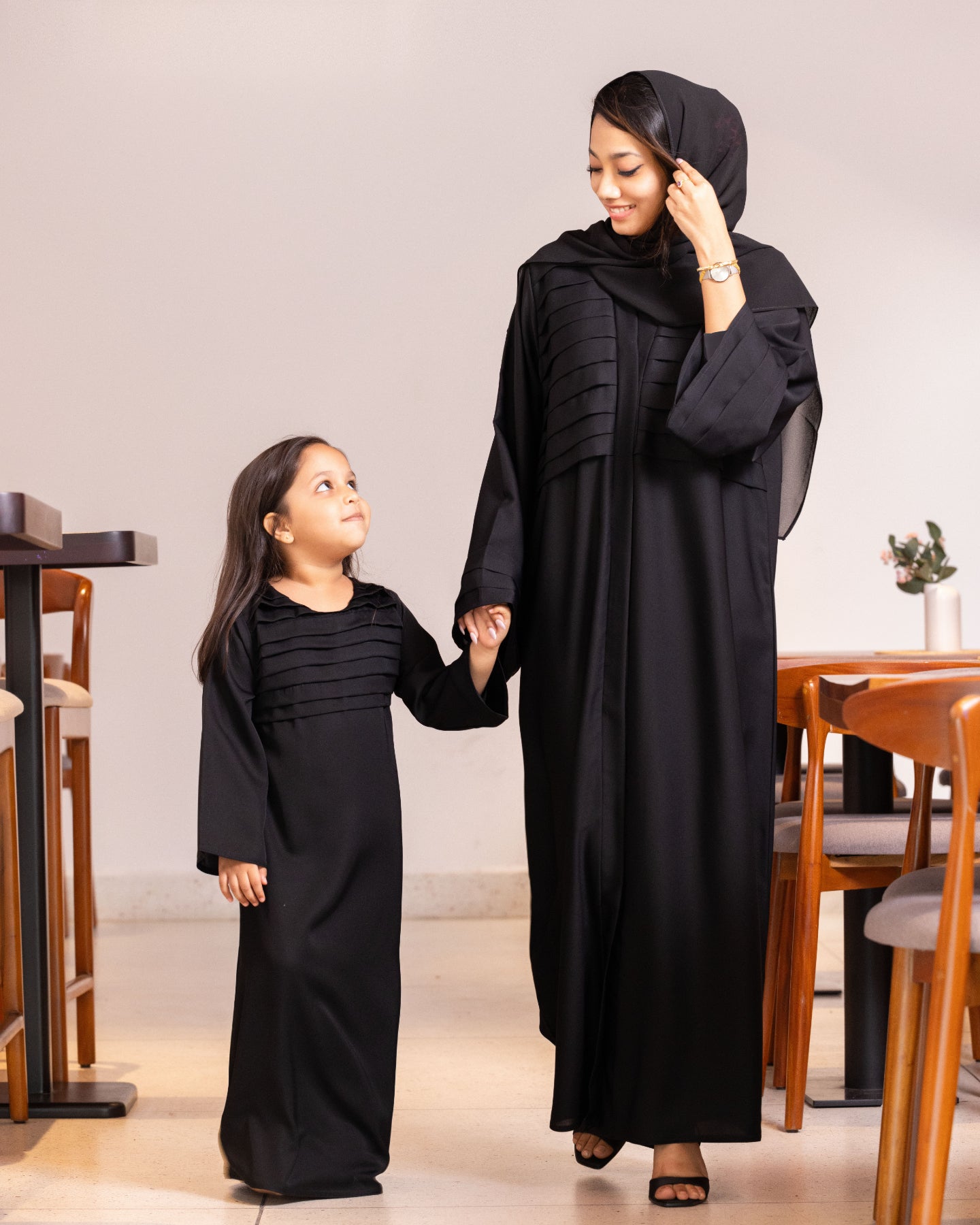 A mother and daughter walking and holding hands while wearing matching black abaya designs with pleated chest details from our latest abaya collection.